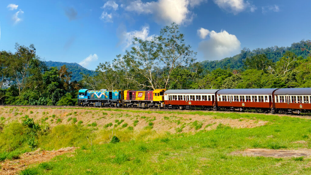 刈り草の残る草地の向こうを走るクランダ観光鉄道の列車。青いアボリジナルアート塗装の機関車と赤黄の機関車が、えんじ色の木造客車を牽引し、背後には熱帯雨林の山と青空が広がる。