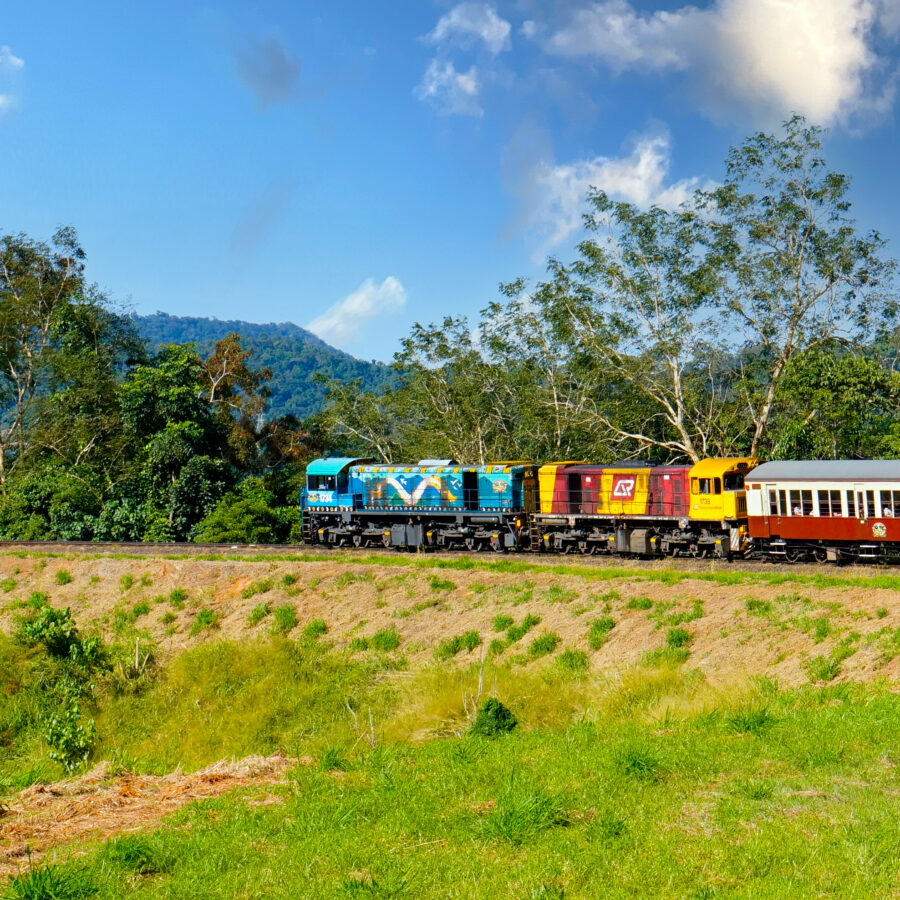 刈り草の残る草地の向こうを走るクランダ観光鉄道の列車。青いアボリジナルアート塗装の機関車と赤黄の機関車が、えんじ色の木造客車を牽引し、背後には熱帯雨林の山と青空が広がる。