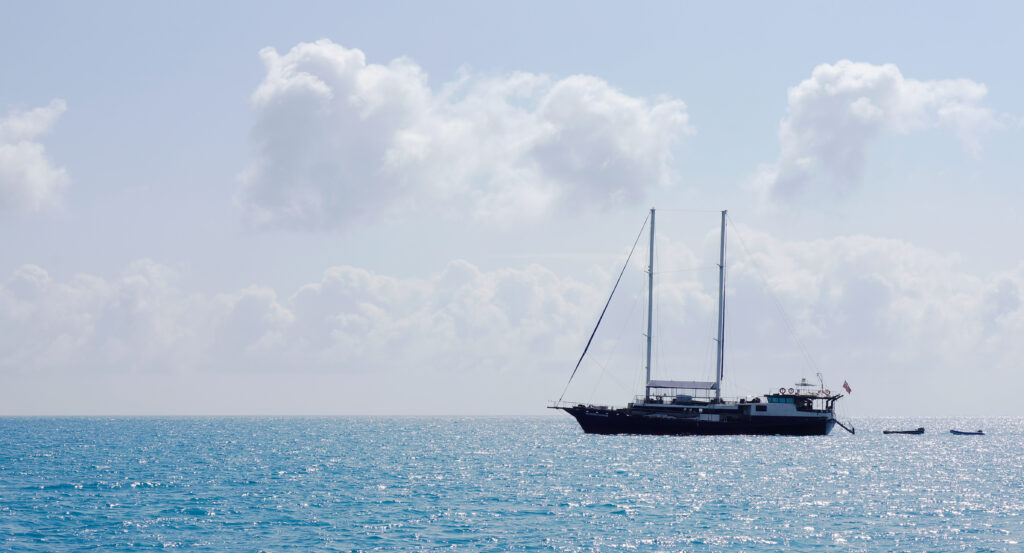 ウィットサンデイ諸島の青い海に停泊する二本マストの帆船「Atlantic Clipper」。白い積雲が広がる空の下、船尾に旗がはためき、右手に小型テンダーが二艘浮かぶ。