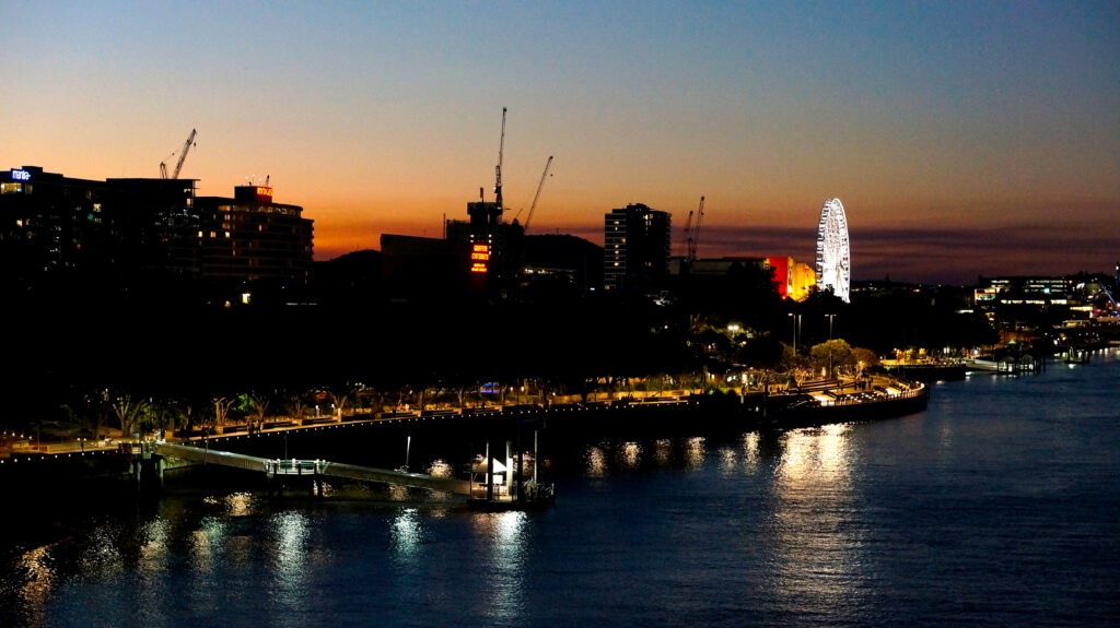 日没後のブリスベン川沿いの夜景。オレンジ色に染まる残照の空を背景に、サウスバンクの観覧車ホイール・オブ・ブリスベンが白く点灯し、遊歩道の灯りが川面に映り込んでいる。