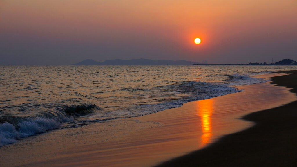 夕暮れの海。オレンジからピンクへと染まる空に太陽が低く輝き、濡れた砂浜と海面に金色の光の帯が長く伸びる。手前には打ち寄せる波、水平線の彼方には岬の稜線のシルエット。タイ・ラヨーンの海岸。