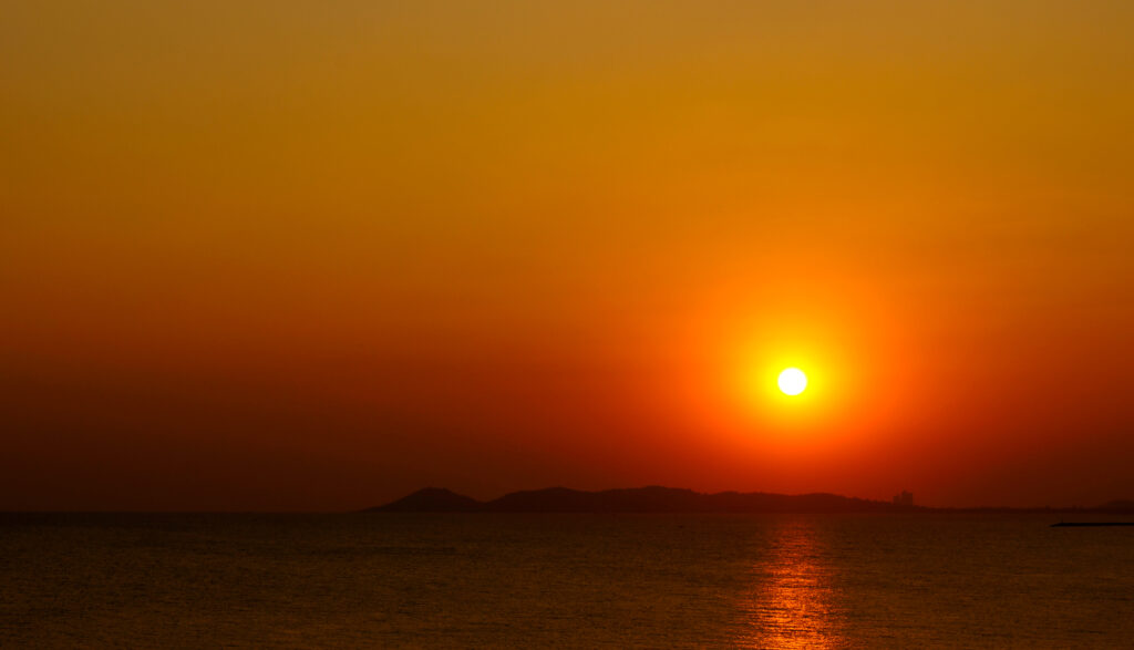 一面オレンジ色に染まった夕空と海。水平線近くに沈みゆく太陽が、穏やかな海面に金色の光を落とす。彼方には島影の稜線が黒いシルエットとなって横たわる。タイ・ラヨーンの海。