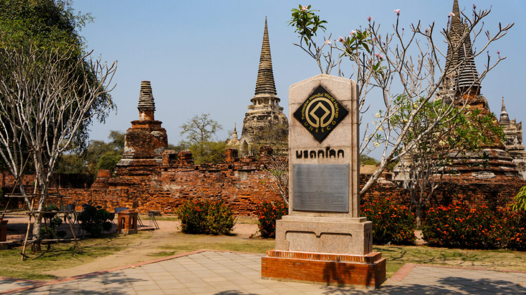タイ・アユタヤ遺跡の世界遺産登録記念碑。「Patrimoine mondial / World Heritage」とUNESCOのロゴ、タイ語で「มรดกโลก（世界遺産）」と刻まれた御影石のモニュメントが立ち、背後にワット・プラシーサンペットの鐘形仏塔（チェディ）とレンガの塀、青空が広がる。手前にはオレンジの花壇とタイル敷きの通路。