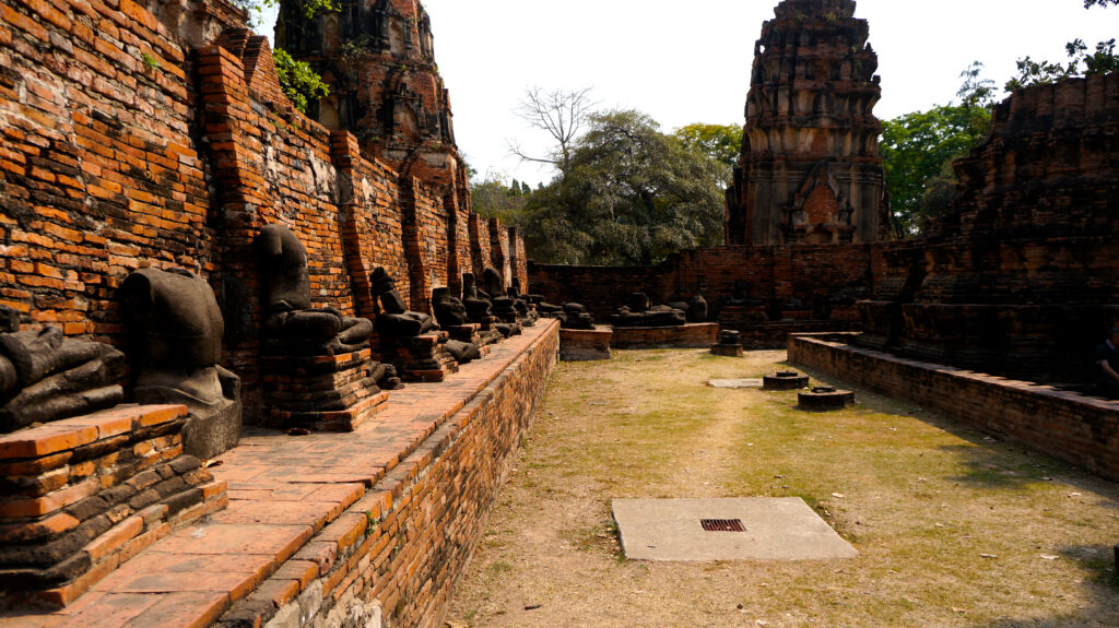 タイ・アユタヤ遺跡の寺院跡。赤レンガの壁に沿って、頭部を失った石造りの仏像が台座の上に一列に並ぶ。奥にはクメール様式の崩れかけた塔(プラーン)がそびえ、境内には乾いた芝生と緑の木々が広がる。